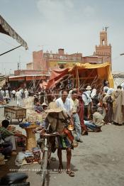 Image du Maroc Professionnelle de  Avec un chapeau de paille sur la tête et poussant son vélo, le traditionnelle vendeur et porteur d'eau dit –Guerrab- n'est pas aussi visible que celui qui est folklorique doté d'un chapeau multicolore, et d'une tenue , chargé de coupelles en cuivres ou de fer blanc qui brille sous les rayons du soleil. Cet authentique vporteur d'eau qui désaltère ceux qui ont soif, traverse l'espace de la Place Jemaa El Fana et ne fait aucunement pas partie des attractions touristiques, au fond le minaret de la Mosquée Kharbouche, Jeudi 19 Mai 1988. (Photo / Abdeljalil Bounhar)